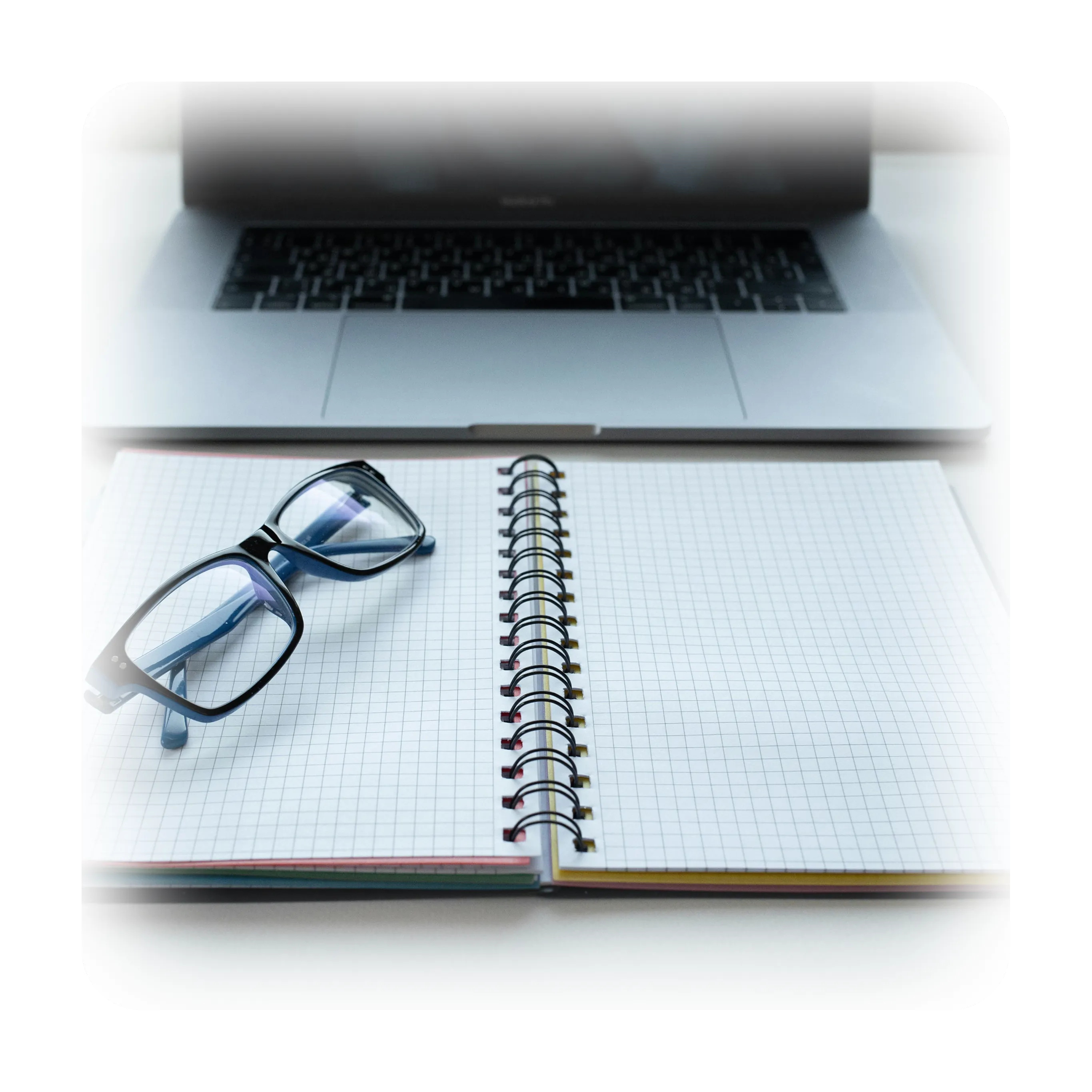 A clean, high-angle shot of an open grid-paper notebook and glasses sitting on a white desk in front of a blurred laptop.